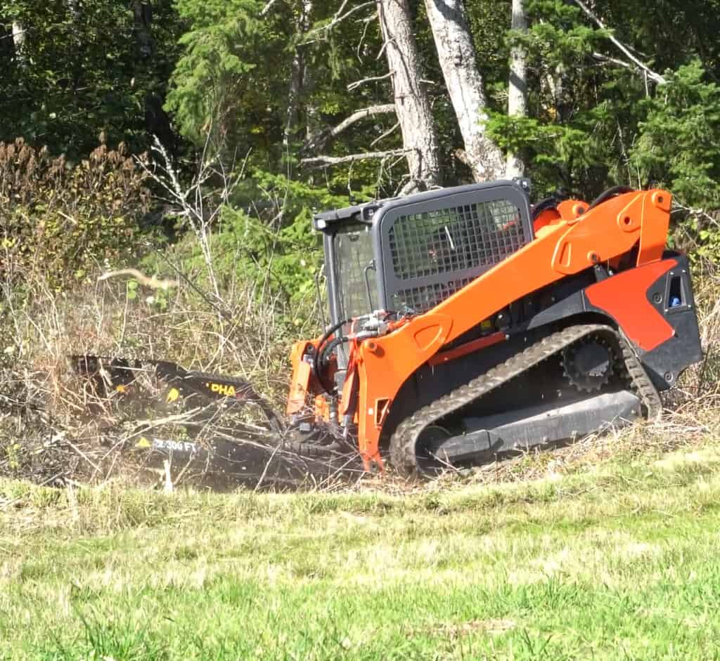 Brush Wolf Alpha skid steer brush cutter side view clearing brush and overgrowth along wooded jobsite edge