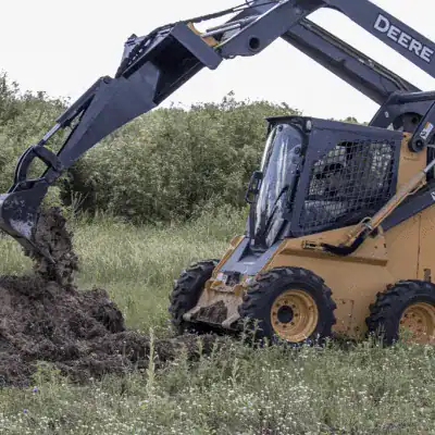 Prime Attachments skid steer backhoe mounted on Deere machine digging brush showing land clearing and trenching use