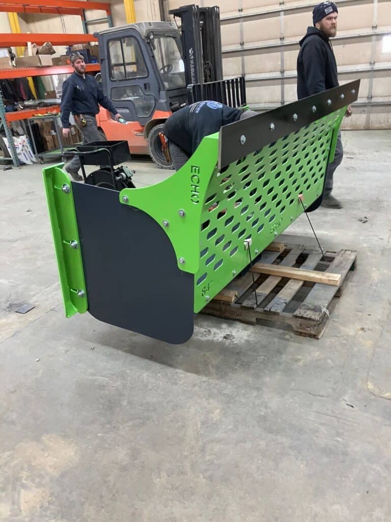 Workers assembling ECHO SSP skid steer snow pusher attachment inside fabrication shop