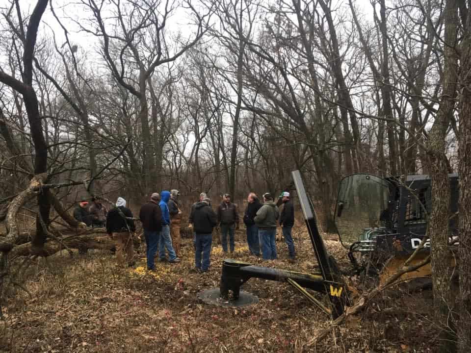 M&M Hydra Saw skid steer attachment demonstration group standing in wooded area