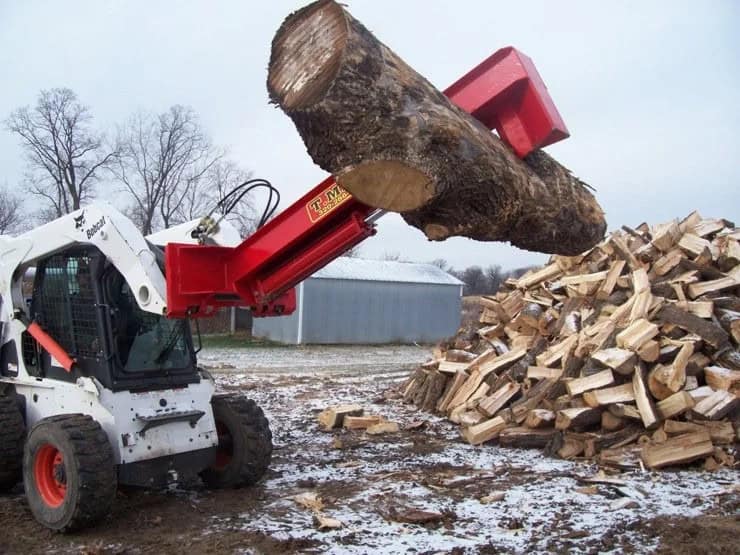 TM Manufacturing Pro 1 skid steer log splitter lifting a large log beside a stacked firewood pile
