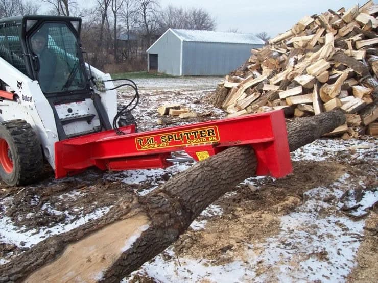 TM Manufacturing Pro 1 skid steer log splitter positioned over a long log beside a firewood pile