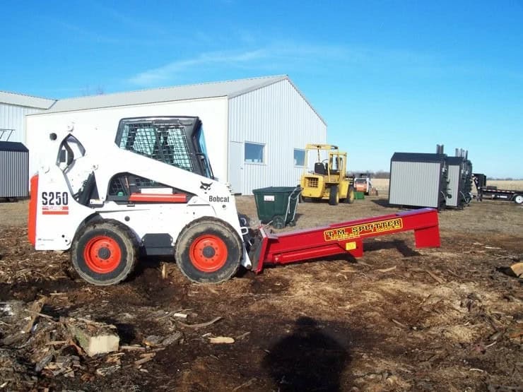 TM Manufacturing Pro 1 skid steer log splitter attached to a Bobcat S250 at a wood processing site