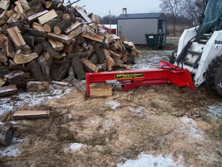 TM Manufacturing Warrior skid steer log splitter working beside a large firewood pile with a Bobcat skid steer