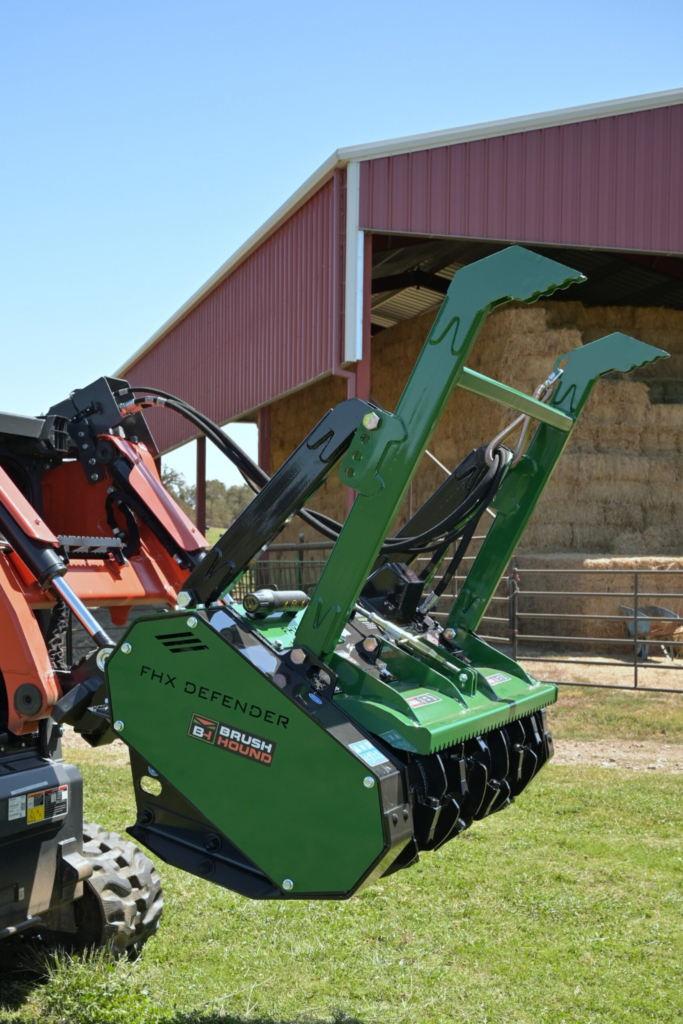 BrushHound FHX66 Defender Forestry Mulcher mounted on a skid steer near a farm building.