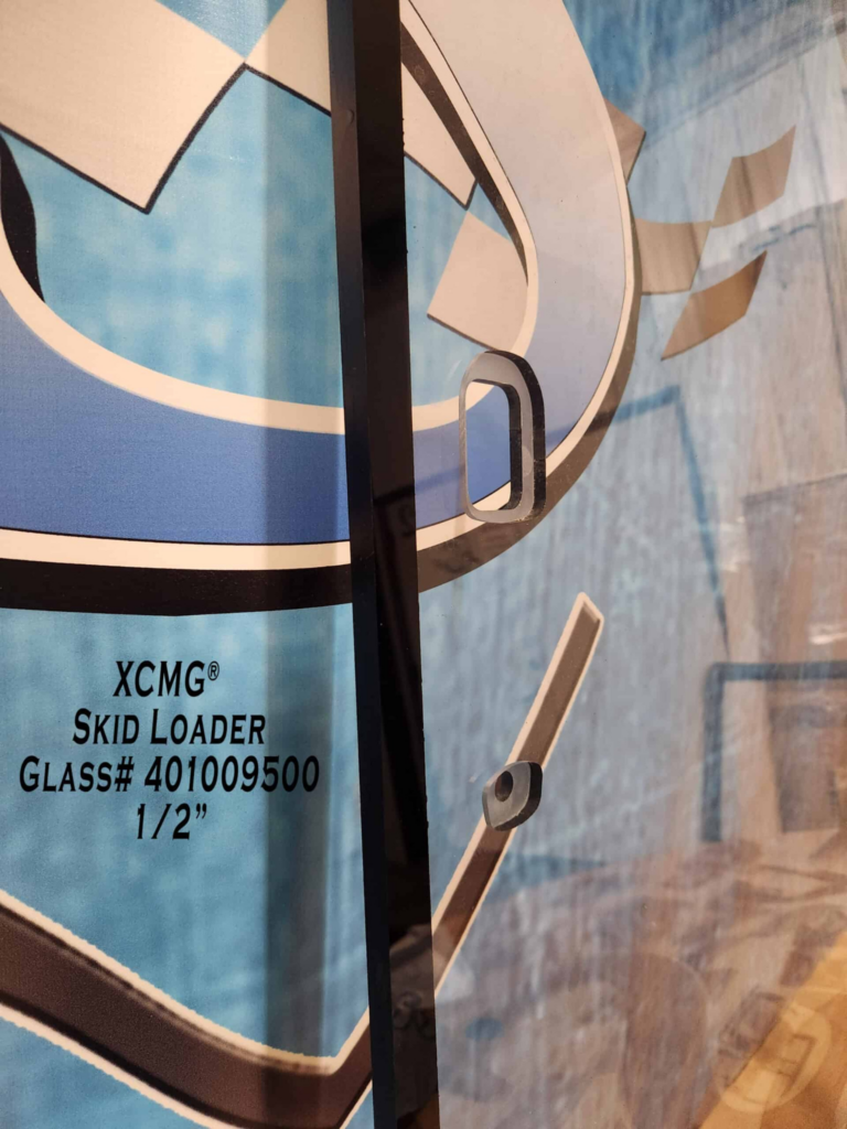 Close-up of SHIELDS XCMG polycarbonate windshield showing the fritted edge and precision-machined mounting holes.