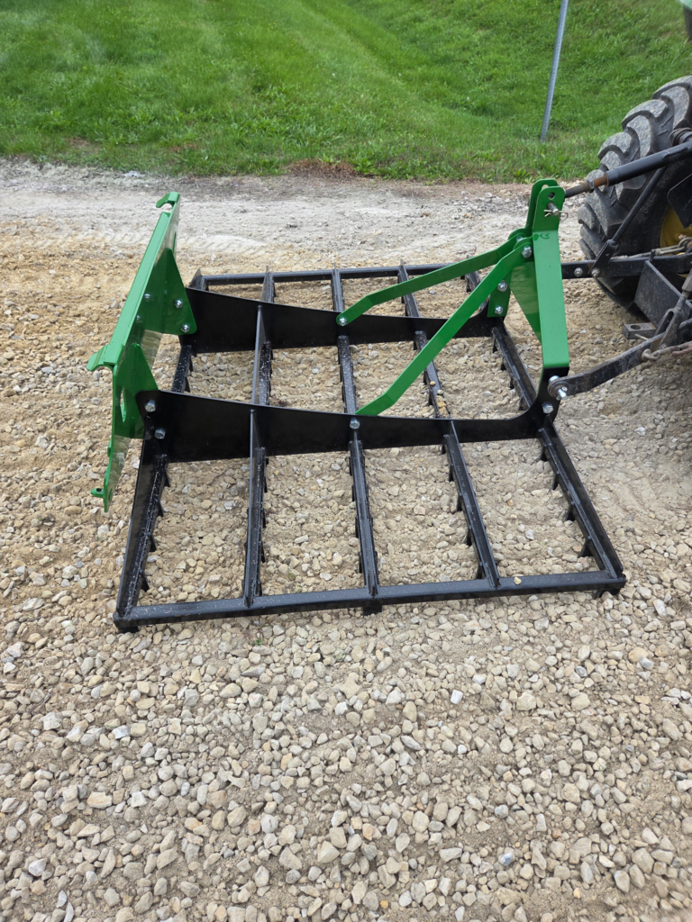Rear angle of the SWAT Metal Works 72" Compact Land Rake attached to a compact tractor for driveway maintenance.