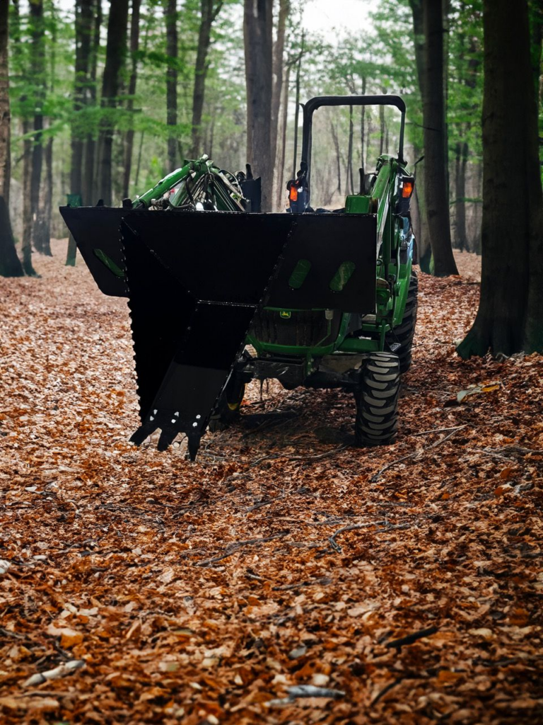 Black SWAT Metal Works Light Duty Stump Buster mounted on a compact tractor in a wooded setting, showing the digging snout and quick-attach plate.