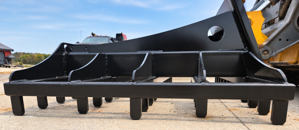 Low close-up of the SWAT Metal Works Skid Steer Land Rake showing the heavy-duty scarifier teeth and reinforced frame